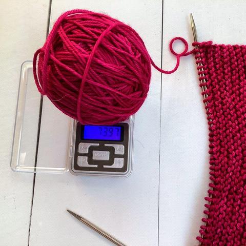 Flat lay view of a ball of dark pink yarn on top of a small scale, and a knitting project featuring garter stitch on a white plank background. The scale indicates that there is 73.97 g yarn left.