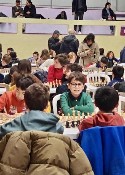 A group of children is engaged in a chess tournament. In the foreground, Elyes, wearing glasses and a green shirt smiles while seated at a table with chess pieces. Other children are focused on their games, and adults are visible in the background. The