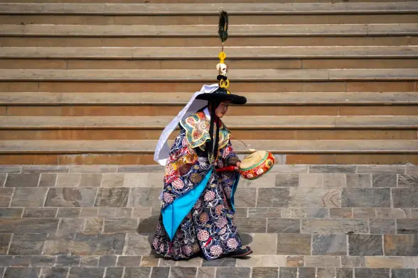 A Sherpa monk dressed in colorful robes and with a vertical head ornament is carrying a small drum.