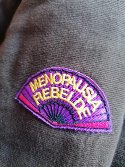Parche de tela para pegar o coser en prendas. Cosido a máquina, es un abanico con las letras menopauisa rebelde en amarillo dorado sobre fondo morado