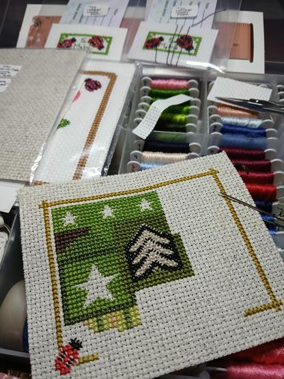 A cross stitch project in progress entitled "Military Patchwork Prayer Cloth" is the main focus in the picture. It has black and tan sergeant stripes in the middle of the piece. Other areas have stars (which will be white once finished) surrounded by olive green colors. Another section has a camouflage section, and still another has light green, tan and darker green alternating stripes. In the background is an embroidery floss bobbin box with several colors of embroidery floss, some cross stitching tools in some of the box's sections and a few other cross stitch projects laying on top of the box that do not yet have all of their backstitch completed.