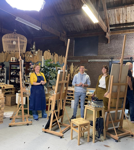 Drie kunstenaars staan elk achter een schildersezel in een nostalgische wijnwinkel en lachen naar de camera