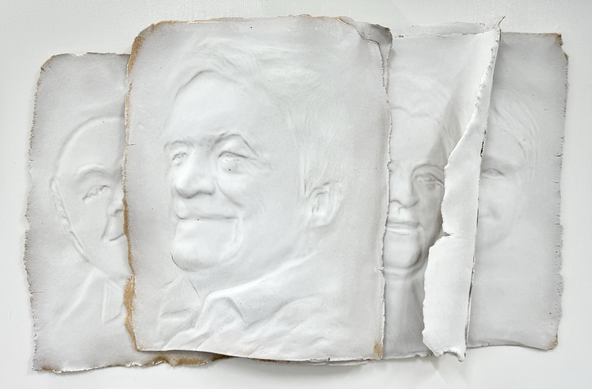 Reliëf van wit geglazuurd keramiek, bestaande uit vier delen die overkomen als papieren half achter elkaar met daarop het reliëf van Johnny Kraaijkamp en op de drie andere delen, zijn vader, zoon en kleinzoon. 