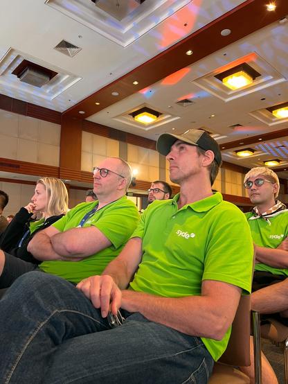 Syde team members in green shirts sit in the audience at WordCamp Asia 2025, attentively listening to a presentation. The conference room features a modern ceiling design with colorful lighting.