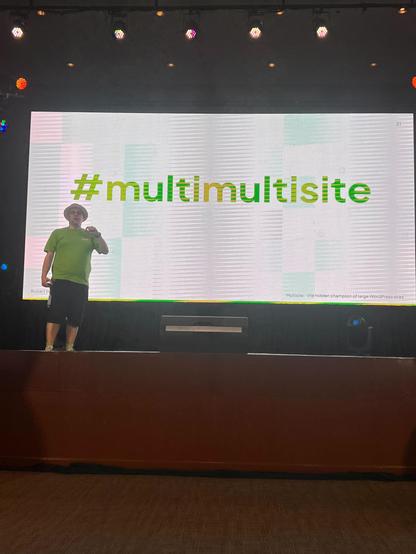 Robert Windisch, a Syde team member, stands on stage at WordCamp Asia 2025, presenting in front of a large screen displaying the hashtag #multimultisite. He is wearing a green t-shirt and a hat.