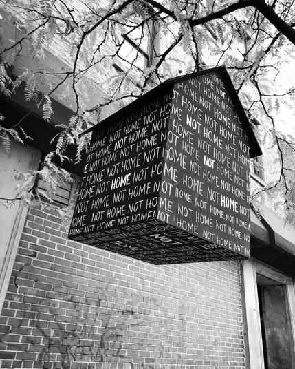 Street art. A wooden house, painted black hangs from a tree branch. The house is completely covered with the words HOME and NOT HOME in plain capital letters. 