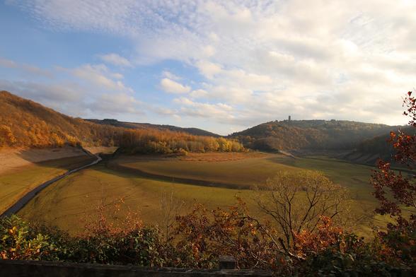 Das Bild zeigt eine Landschaftsaufnahme. Man sieht ein Flusstal, das grün, aber seltsam leer erscheint. Es handelt sich um die Urfttalsperre, die zu Inspketionsarbeiten leer gelassen wurde. Es scheint die Sonne, die schon tief steht, aber nicht zu sehen ist. Man erkennt nur die langen Schatten. Der Himmel ist blau mit leichter Bewölkung. In der Ferne ist der Turm der ehemaligen NS-Ordensburg Vogelsang zu sehen.