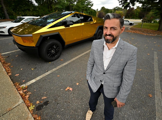 "A Worchester man chose to color his Cybertruck shiny gold wrap.
He also chose to wear light beige loafers with his black pants and light grey, glen plaid jacket, for his 15 minutes of professional- photography-subject fame with a local news crew."

Additionally, his sartorial choices make him appear as though he might spike your drinks and have a massive crypto portfolio, that he absolutely has to tell you about. 

 His smile only confirms this assumption. 