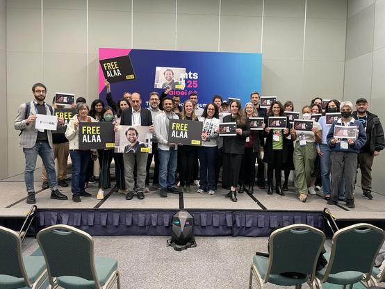 A group of people on a stage stand in front of a RightsCon 25 banner. They hold photos of Alaa Abd El Fattah and signs that say FREE ALAA.