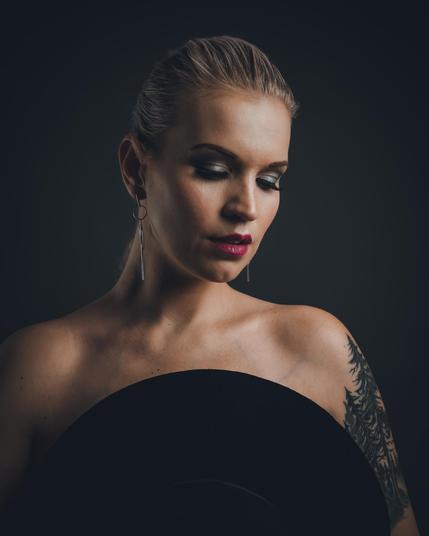 A close-up portrait of a woman looking upward towards the light. Her hand is resting behind her head, creating a strong yet elegant pose. The lighting emphasizes her glowing skin and sharp facial features against a dark studio background.