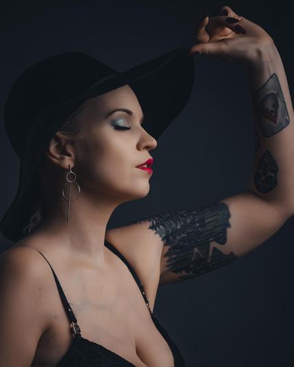 A moody portrait of a woman in studio lighting. She gazes downward, with dramatic lighting highlighting her features. The image includes a black hat, silver minimalist earrings, and a tattooed shoulder. The background is dark and neutral.