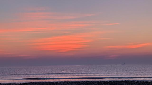 Historical Documentation in the form of a photograph of a beautiful sunrise over the ocean. The sky is filled with vibrant orange and pink hues, with the sun setting on the horizon and casting a warm glow over the water. The low clouds glowed with the first rays of the morning sun. The water is calm and reflects the colors of the sky, creating a peaceful and serene atmosphere. In the distance, there is a small boat visible on the water's surface. The beach is covered in small pebbles, and the horizon line is visible in the distance. The overall mood of the image is one of calmness and tranquility.