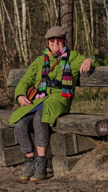 Eine Frau sitzt auf einer Bank. Es ist sonnig. Bekleidet ist die Frau mit einem grünen Mantel. Ein bunter langer Schal und eine Schirmmütze ergänzen die Bekleidung wie auch Wanderschuhe. Eine Kamera und eine kleine Tasche hängen mit Riemen um den Hals der Person. Die Brille hat verdunkelte Gläser. Die dunkelgraue Hose lässt nackte Beine erkennen.