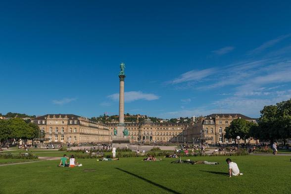 Sommerliche Szene um das neue Schloss in Stuttgart mit Jubiläumssäule. Vor dem Schloss ist eine grüne Wiese zu sehen, auf der Menschen sitzen.