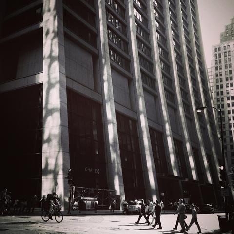 Black and white photo of the base of the Chase Tower on Dearborn and Washington. Dappled sunlight reflects on the pedestrians cars and bicyclists at street level. 