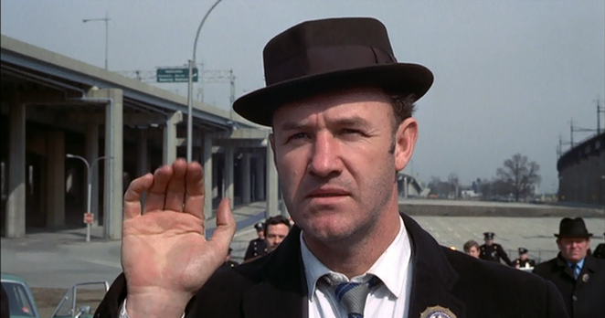 Eine Szene aus dem Film "French Connection". Gene Hackman im Vordergrund, er hebt seine rechte Hand zu einem Abschiedsgruß. Hinter ihm weitere Menschen, hinten links ist ein Brückenbauwerk zu sehen. Auf Gene Hackmans Kopf ist ein Hut mit Krempe.