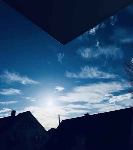 Motiv:
Das Bild zeigt eine städtische Wohngegend mit mehreren Gebäuden im unteren Bereich. Der Himmel nimmt den Großteil des Bildes ein. Die Sonne steht tief und ist leicht von Wolken verdeckt. Eine Dachkante ragt von oben ins Bild hinein.

Stimmung & Wirkung:
Das Bild zeigt eine klare Himmelsszene mit starkem Kontrast zwischen Licht und Schatten. Die Silhouetten der Gebäude betonen die Helligkeit des Himmels.

Technische Details:
Farbe: Dominierende Blautöne mit starken Kontrasten zwischen Himmel, Wolken und dunklen Gebäuden.
Schärfentiefe: Der Himmel und die Wolken sind scharf, während die Gebäude im Schatten wenig Details zeigen.
Lichtverhältnisse: Starkes Gegenlicht durch die Sonne mit ausgeprägten Kontrasten zwischen hellen und dunklen Bildbereichen.