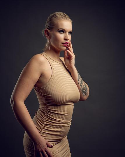 A professional studio portrait of a blonde woman wearing a form-fitting beige dress, posing elegantly with her hand near her lips against a dark background. The lighting highlights her natural curves and tattoos, emphasizing a confident and stylish expression.