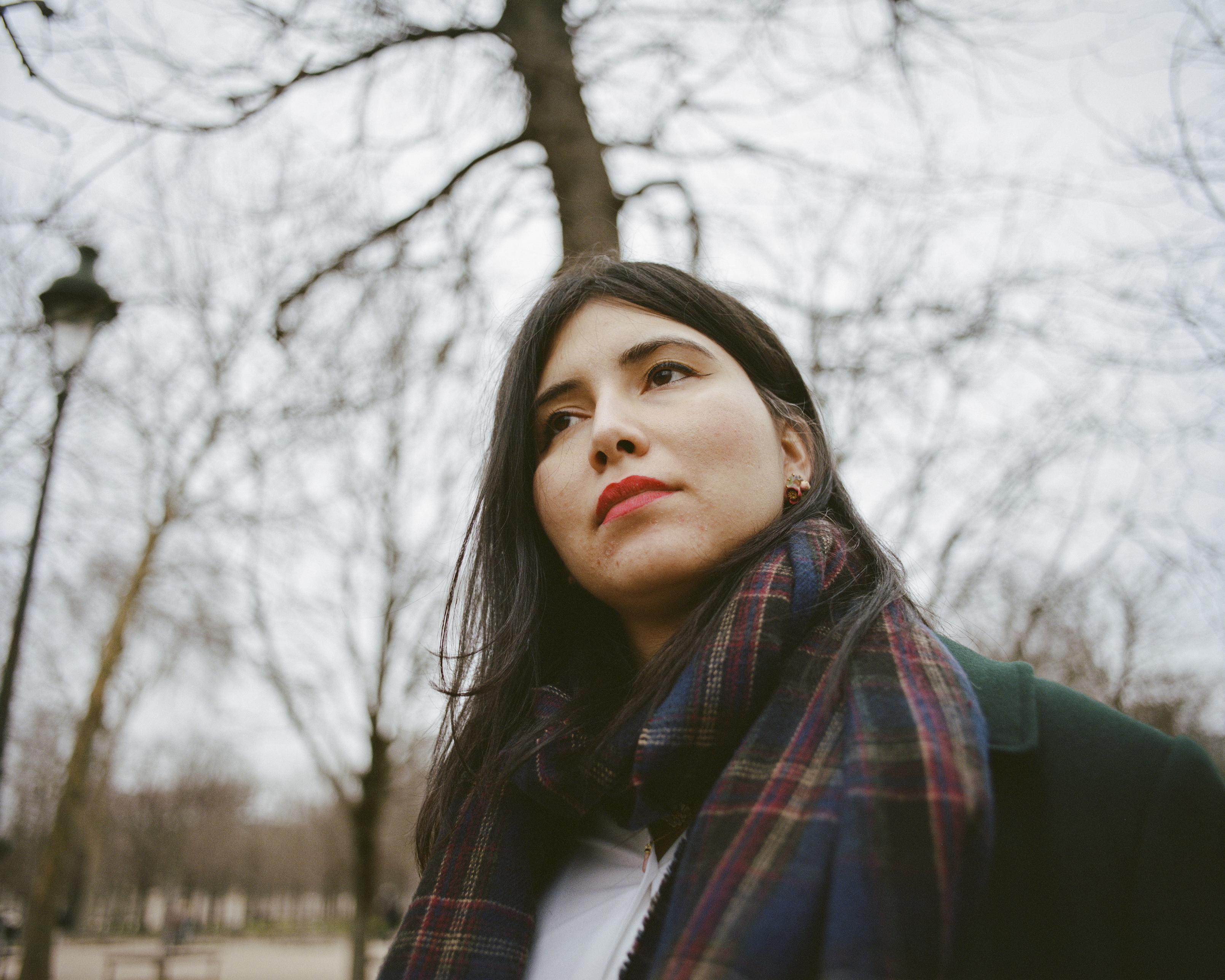 A beautiful woman with dark, straightened hair and red lipstick, wearing red floral earrings, a navy tartan scarf and a green woolen coat. In the background, barren trees and a lamp post.