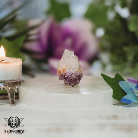 One specimen of the small "Sugar Amethyst Cluster" product is arranged on a selenite disc next to a burning tealight candle. The crystal has clear quartz and amethyst points with smaller crystals growing on the larger ones, giving a sugared look. Presented by Inked Goddess Creations.