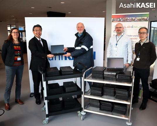5 Personen, eine Mitarbeiterin und 3 Mitarbeiter von Asahi Kazei und ein Labdoo-Helfer stehen hinter 2 Rollwagen voller Stapel mit Laptops. Im Hintergrund 2 Poster von Asahi Kazei und Labdoo. Alle schauen in die Kamera. Der Geschäftsführer der Firma und der Labdoo-Helfer halten einen aufgeklappten Laptop zwischen sich.