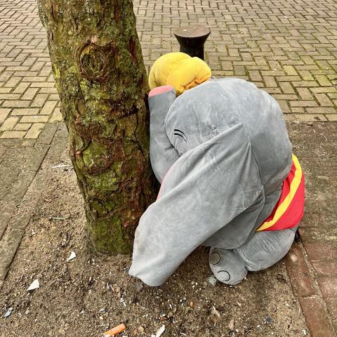 A large plush toy elephant rests with its trunk against a tree, with its head bowed down. It’s Dumbo. The setting is an outdoor space with a brick walkway and some debris on the ground.