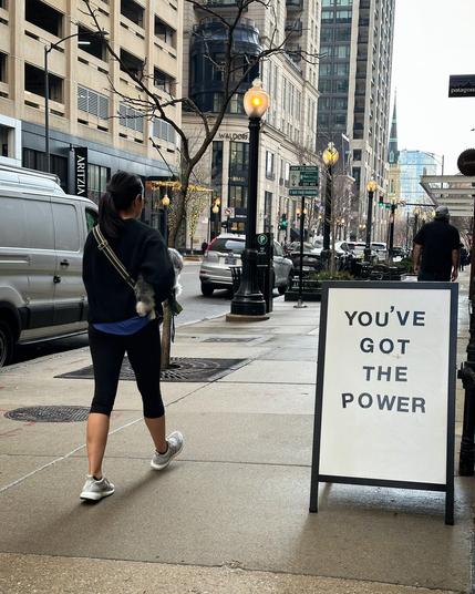 A woman walks past a sandwich board in the sidewalk that says “YOU’VE GOT THE POWER.” She is walking through the Gold Coast, arguably the most expensive residential and shopping district in Chicago 