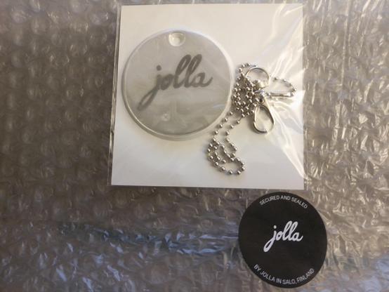 Top left a reflctive washer with the Jolla logo with chain and zipclip on top of white square paper. Bottom right a round Jolla sticker keeping the bubble plastic wrapping in place.
White text on black. Center: Jolla logo 
Surrounded with text alog the rim.
Above: Secured and sealed
Below: By Jolla in Salo, Finland