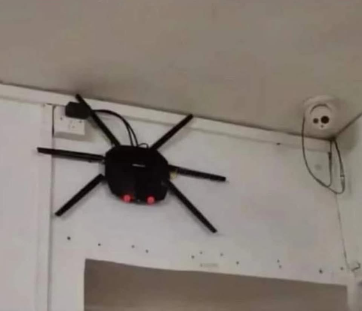 A black, spider-like router with six legs and red lights is mounted on a white wall near a power outlet. A round, white security camera is mounted higher on the same wall to the right.