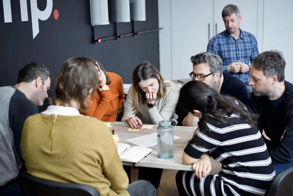 A group of people gathered around a table, engaged in a collaborative workshop. They are looking at sticky notes and papers with focused expressions. A facilitator stands in the background, observing the discussion.