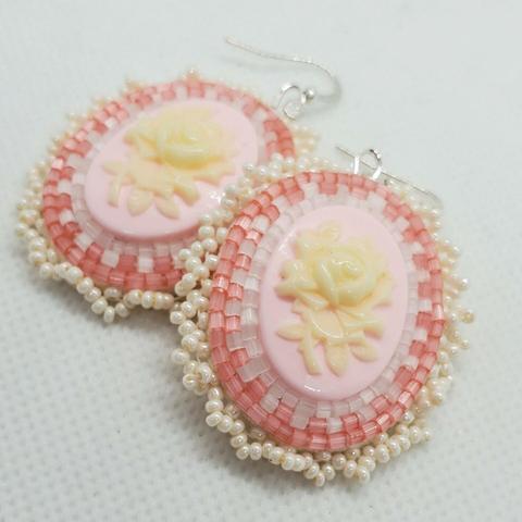 a pair of beaded oval-shaped earrings. the centerpiece is a delicate cream colored rose on a pale pink background. the border beads are in pink and clear with a frilly cream colored edge