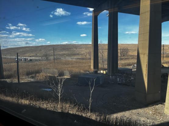 Out the window of a Morris and Essex Line New Jersey Transit west-bound train can be seen pillars supporting the I-95 bridge across the Passaic River. Shipping containers lie in its shadow. Beyond is the hump of Landfill 1-D, covered in straw-colored winter grass. A blue sky with scattered clouds tops it.