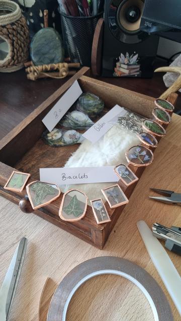English Stained glass pendants with a copper line around them. They contain ivy leaves, ferns and different blue flowers. They are arranged around a small drawer. A label reads "bracelets". Other pieces of glass containing flowers and leaves can be seen further on. Work tools can be seen in the foreground.

Français Pendentifs en vitrail entourés d'une ligne de cuivre. Ils contiennent des feuilles de lierre, des fougères et différentes fleurs bleues. Ils sont disposés tout autour d'un petit tiroir. Il y a une étiquette où il est écrit "bracelets". On aperçoit d'autres morceaux de verre contenant des fleurs et des feuilles plus loin. On aperçoit au 1er plan les outils de travail.