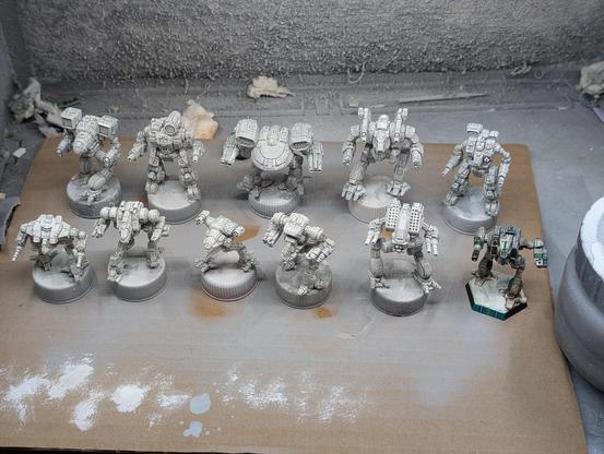 Ten mech miniatures painted white along side a finished miniature painted as Clan Jade Falcon Gamma Galaxy in winter camouflage.