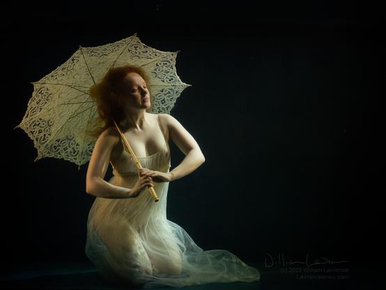 Model Ivory Flame poses in a sheer gown and a white parasol, kneeling underwater.  She holds the parasol over her shoulder, and her hair and the hem of the gown drift in the water.