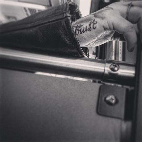 Black and white photo of a hand resting on the divider between the door and the seats on the subway. A tattoo reading “Trust” in script letter is visible on the side of the palm. 