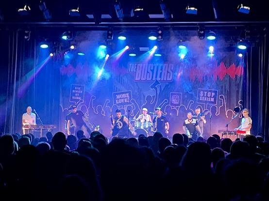 A live band performs on stage with colorful lights, surrounded by a cheering crowd. The backdrop features the band's name, "The Busters," along with silhouettes of people holding protest signs. The musicians play various instruments, creating an energetic atmosphere.