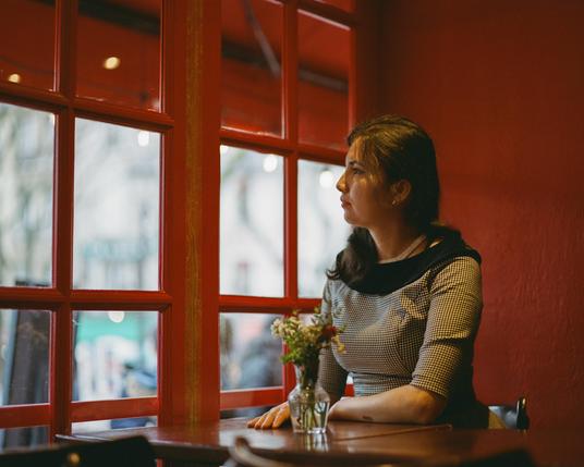 A beautiful woman with dark hair, wearing a black and white dress, is sitting at a wooden table in front of a red paned window and a red stone wall. In front of her is a glass vase with flowers.