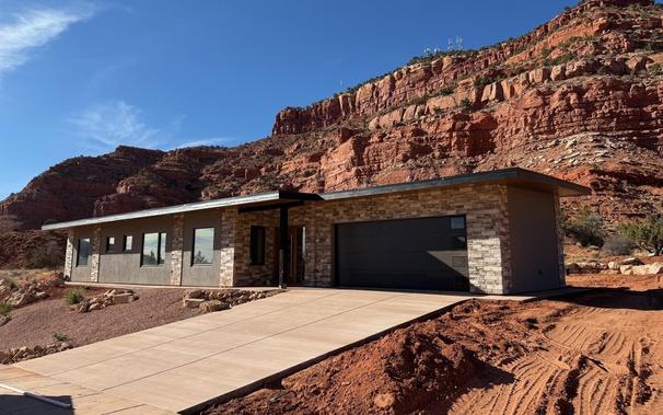 House with a new driveway in front of beautiful red cliffs. Part of the xeriscape yard in front in complete, part of a rock wall can be seen behind the house.