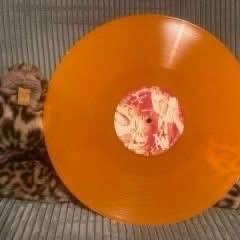 Eine orangefarbene Schallplatte ruht auf einer strukturierten, gemusterten Oberfläche. Das mittlere Etikett hat ein farbenfrohes Design.