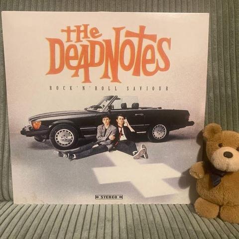 Ein Albumcover von "The Deadnotes" mit dem Titel "Rock 'n' Roll Saviour", mit zwei Männern, die neben einem schwarzen Cabrio sitzen. Ein kleiner Teddybär mit einer blauen Fliege wird neben dem Albumcover platziert.