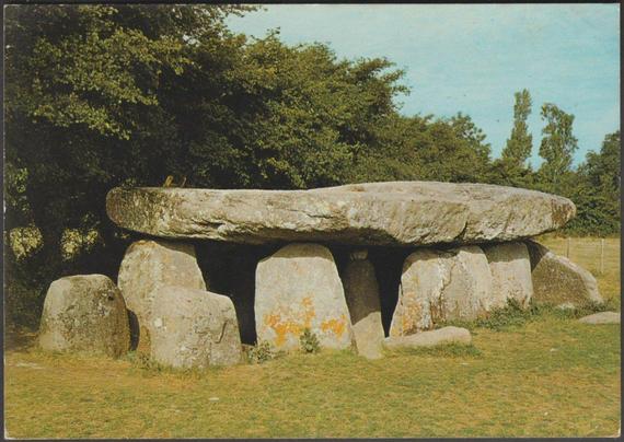 A printed semi-modern postcard showing the Pierre Couverte a la Frébouchère or Dolmen de la Frébouchère near Le Bernard in Vendée, Pays de la Loire. This Angevin-type dolmen is 7.2 m by 3.5 m or 23.5 ft by 11.5 ft, and 2 m or 6.5 ft high and has a massive single capstone, estimated to weigh above 80 tonnes, covering the whole length. The prehistoric burial chamber sits in a field of short green grass with a long line of trees immediately behind it. The sky is blue and cloudless.