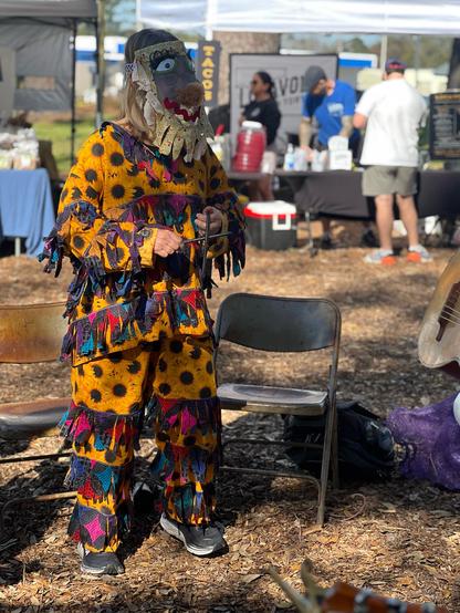 A musician dressed in a traditional courir de Mardi Gras costume plays a T-fer, standing in front of two folding chairs with a farmer’s market behind them. 