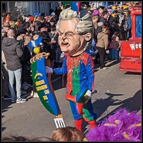 A person in a carnaval parade wearing a mask depicting Dutch far right politician Geert Wilders. He's wearing a joker suit, scepter and crown and a sign with the text  "Alle bitjes helpe. Närrische cultuurstad/länd 2025"