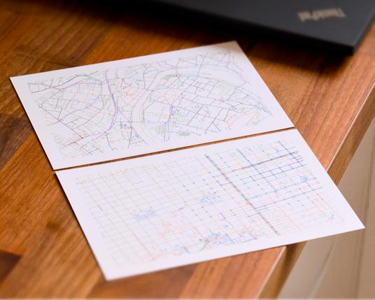 Two postcards with multicolored lines on a wooden desk: One with a grid-like design, the other looking a bit more organic. A ThinkPad in the background.