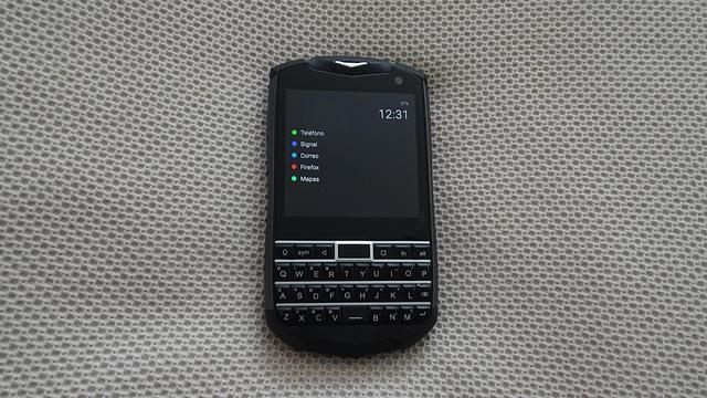 Un teléfono móvil pequeño, con teclado físico (Unihertz Titan Pocket), sobre una superficie de tela. En la pantalla, con fondo negro, la hora, el porcentaje de batería y un listado en texto de cinco aplicaciones (Teléfono, Signal, Correo, Firefox, Mapas). Delante del nombre de cada aplicación hay un círculo en color.