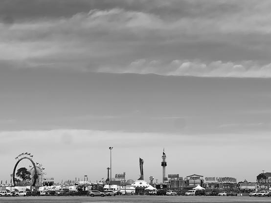 Black and white photo. A carnival scene is far in the distance low on the horizon. A Ferris Wheel is on the left. You can see various tents and rides as well. 