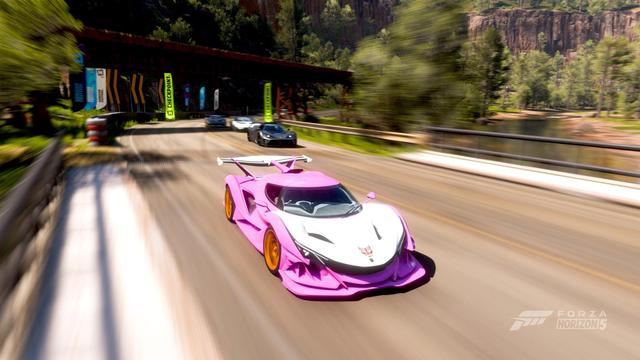 An Apolo IE in Forza Horizon 5 in a race, crossing the bridge by Copper Canyon. The car is sporting a pink and white livery based on the colour scheme of the Gen 1 Transformer Arcee. Where the Autobot logo would normally be, is the stylized logo of the band Cybertronic Spree.