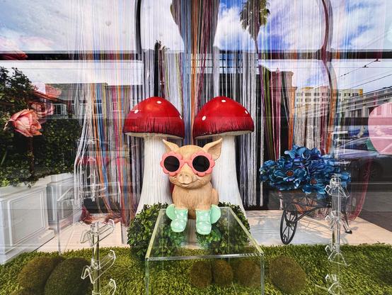 A very fun photo of an adorable pink pig with green boots statue standing in front of two red-topped mushroom plushies. The photo was taken into a storefront window, showing the insides of the store but also reflections from across the street—making the scene almost psychedelic-looking. Everywhere you look it’s filled with all sorts of fantastical objects green mossy grass? A bicycle cart filled with blue flowers, green hedges with big pink roses, multi-colored beaded curtains, and in the background some tall San Francisco-style multi-story residences and shops with palm trees and blue skies with big puffy white clouds. It’s a visual kaleidoscope of colors and themes. It makes me very happy. This was taken on Doc Pop’s recent #pixelwalk in the Mission District of San Francisco on March 1, 1025.