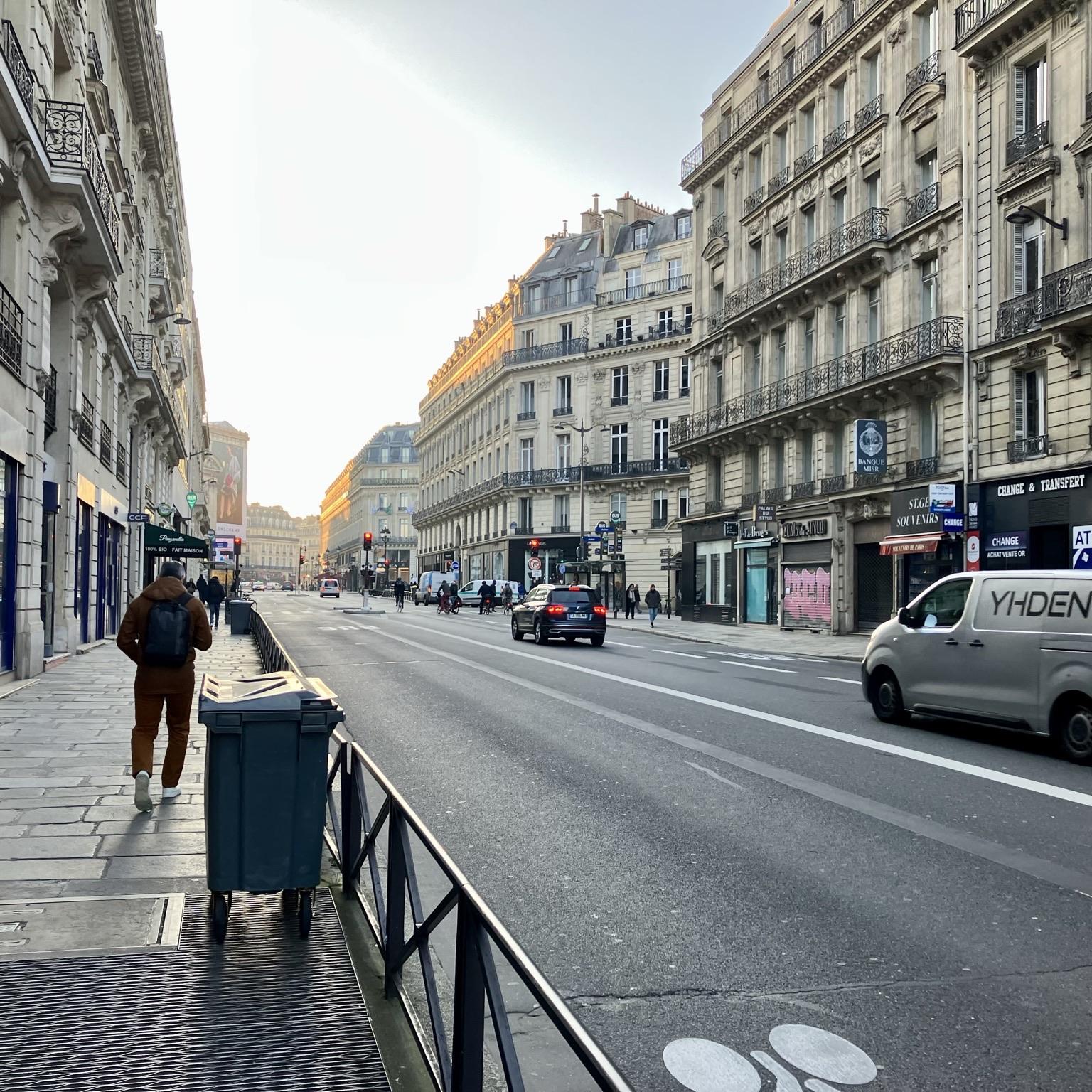 Paris street scene with cars and pedestrians in the morning light, with sunrise glinting off of buildings.